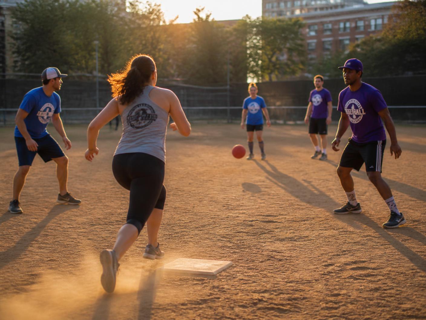 Adult kickball players on a dusty park diamond at golden hour as a runner sprints to first base
