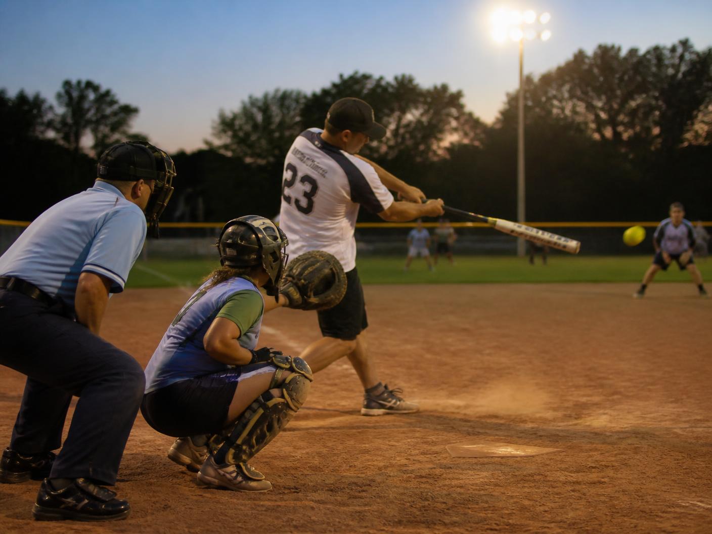 Coed softball batter making contact at dusk with catcher and umpire behind the plate