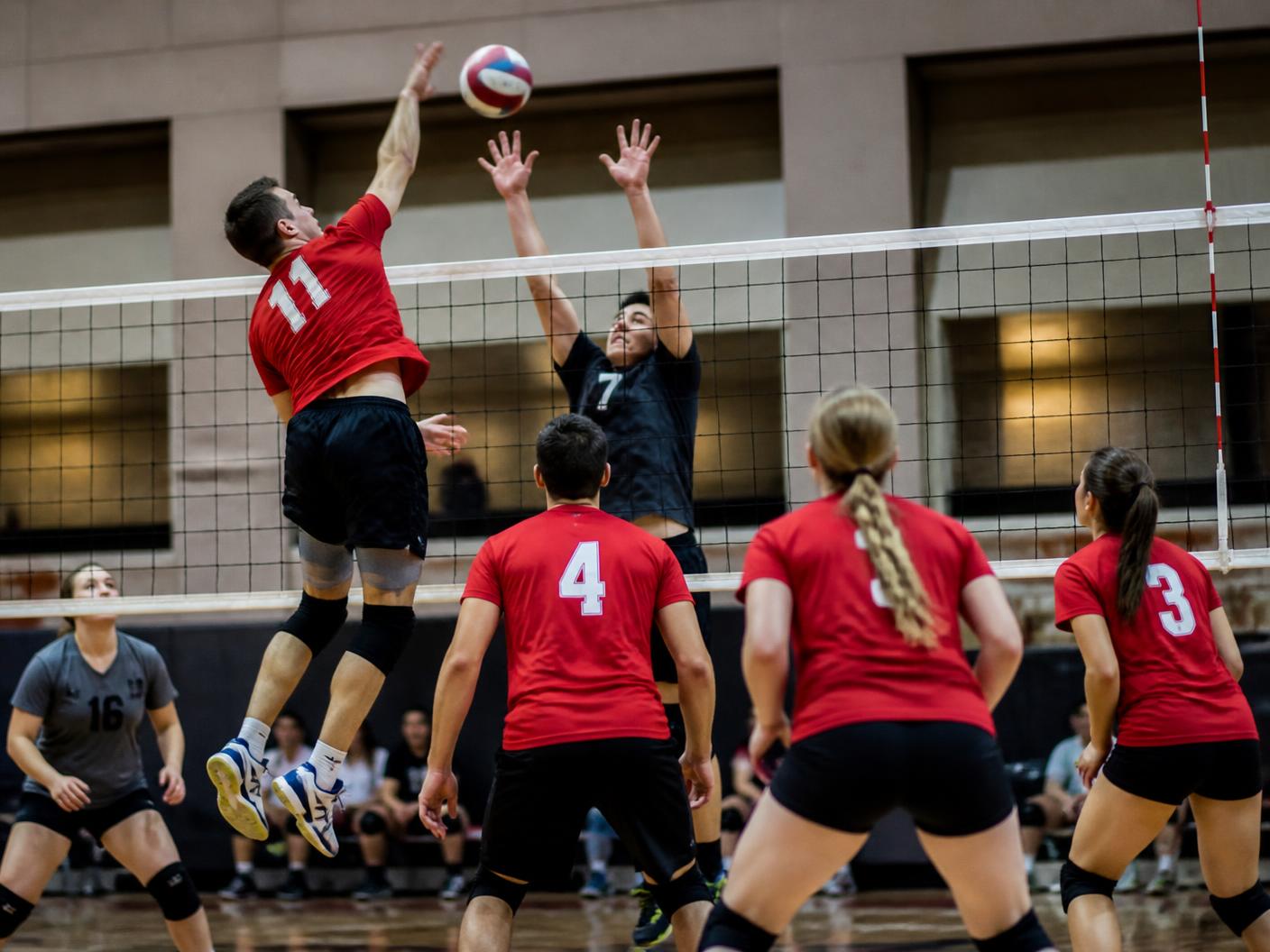 Indoor coed volleyball players at the net, one leaping to spike while blockers reach up