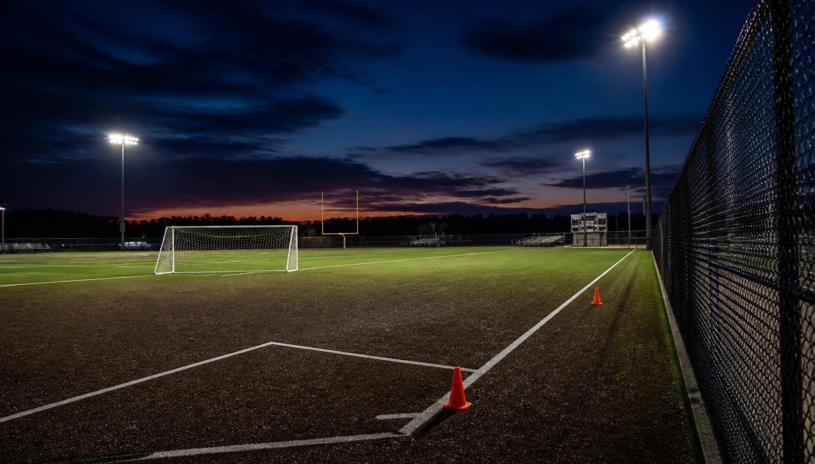 Empty recreational field at dusk with chalked baselines, a soccer goal and orange cones under stadium lights
